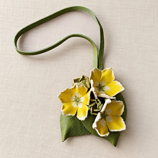 Hand-painted yellow leather floral bag charm with three sculpted flowers, olive-green leaves, and a long leather loop on ivory linen.