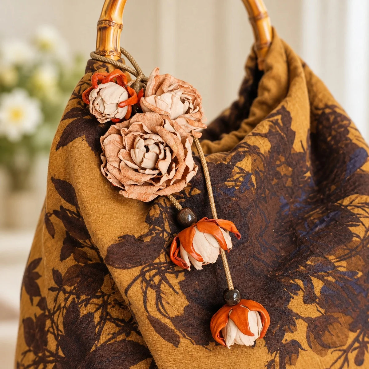 Close-up of beige leather rose bag charm hanging from a bamboo handle on a brown patterned handbag, with sculpted blossoms and cream floral buds.