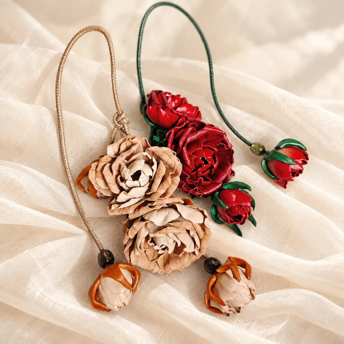 Beige and crimson red handcrafted leather rose bag charms displayed together on ivory fabric, showing contrasting floral colors and hanging cords.