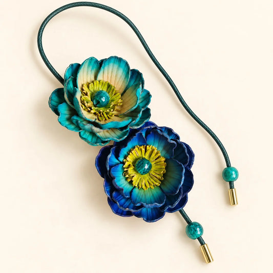 Handcrafted double bloom leather bag charm with teal and royal blue sculpted flowers, green cord and decorative bead ends on an ivory background.