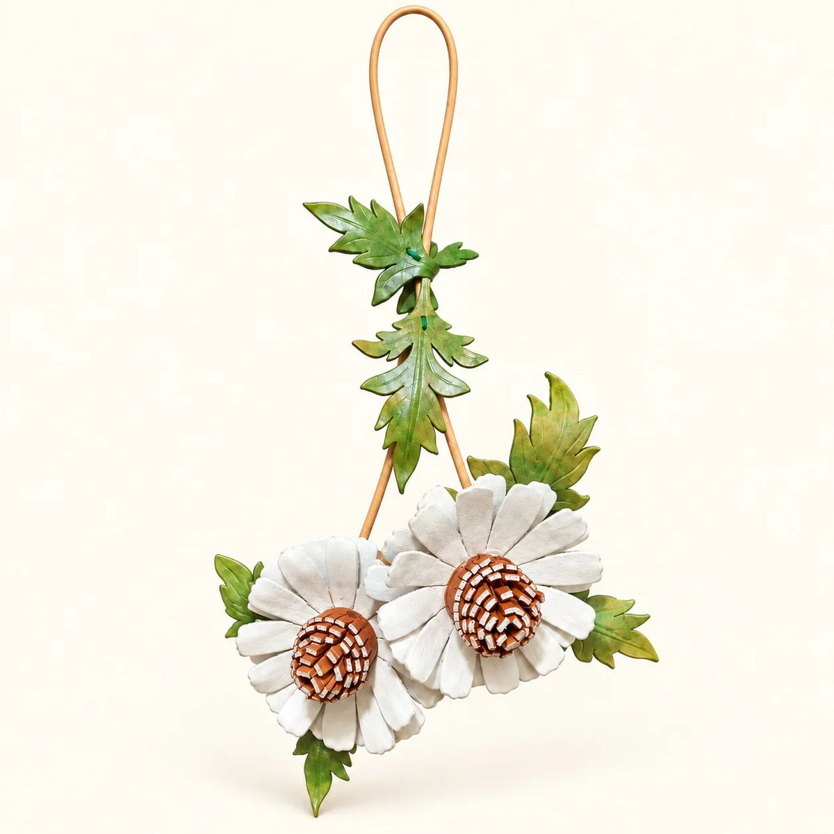 White double daisy leather bag charm with brown textured centers, green lobed leaves and a tan hanging loop on an ivory background.