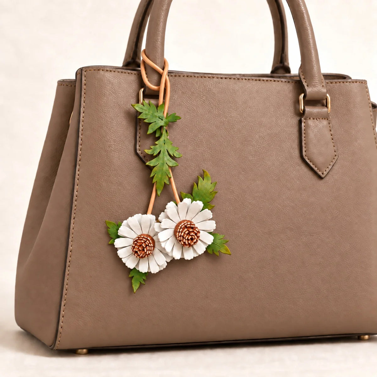 Close-up of a white double daisy leather bag charm attached to a warm taupe handbag handle with a tan loop-through cord.