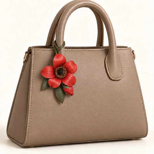 Red kapok flower bag charm attached to the right handle of a cognac satchel with an olive green loop-through strap.