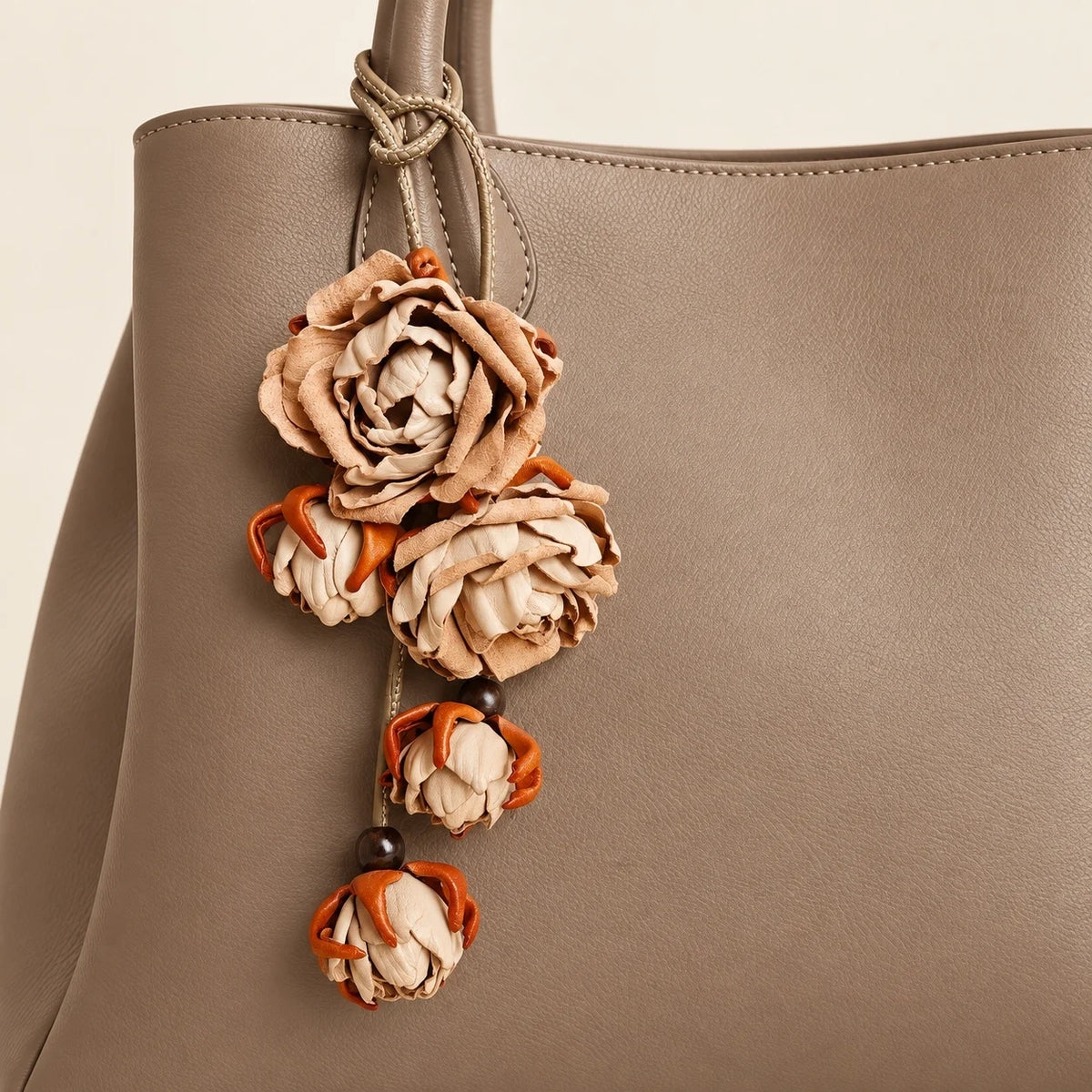Beige sculpted leather rose bag charm secured to a warm taupe tote handle, showing layered petals, dangling buds, and a looped leather cord.