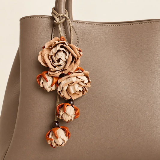 Beige sculpted leather rose bag charm secured to a warm taupe tote handle, showing layered petals, dangling buds, and a looped leather cord.