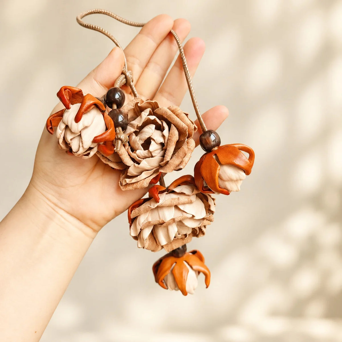 Hand holding a beige handcrafted leather floral bag charm with layered roses, orange-accented buds, dark decorative beads, and a long cord.