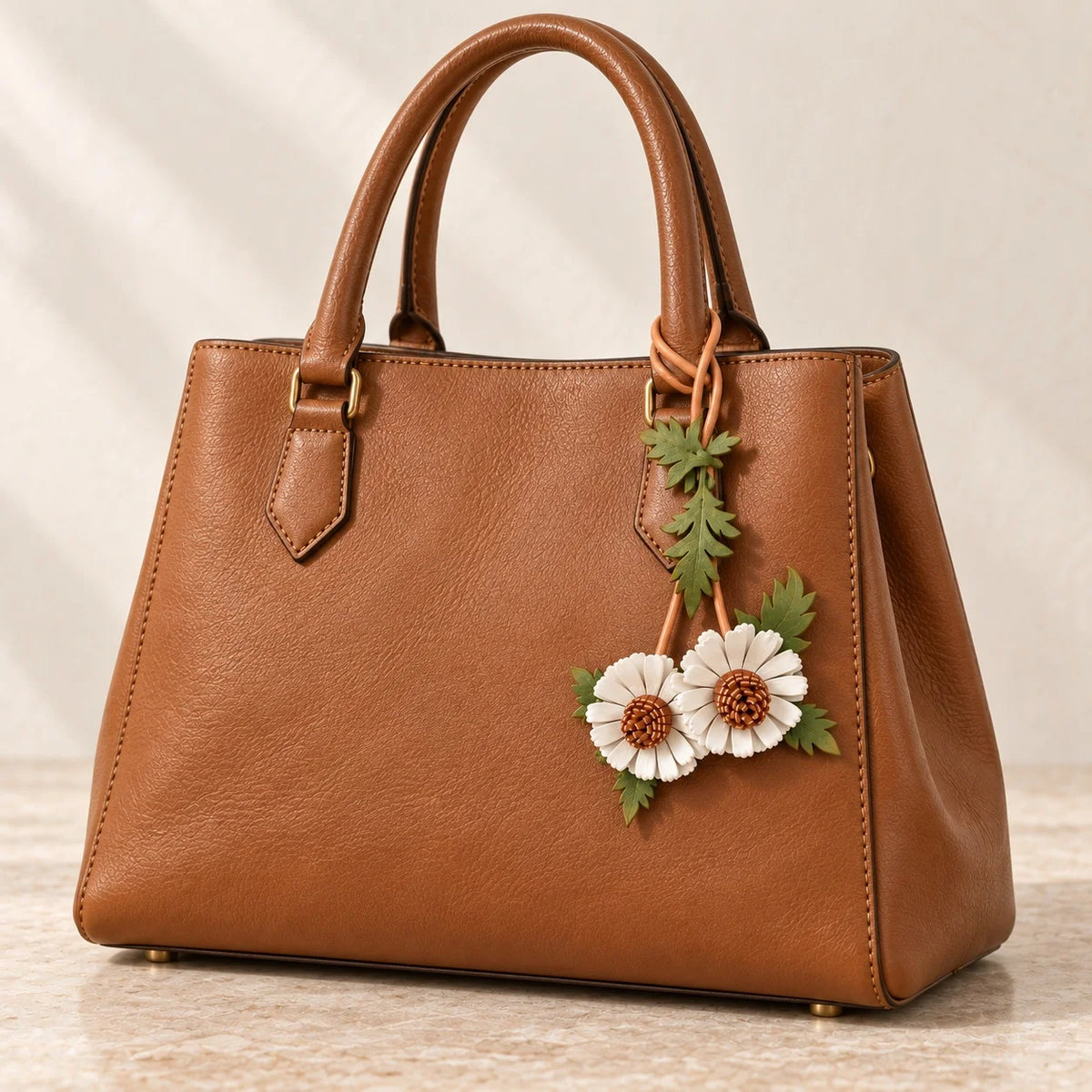 Cognac structured handbag styled with a white double daisy leather bag charm on the right handle, with brown centers and green leaves.