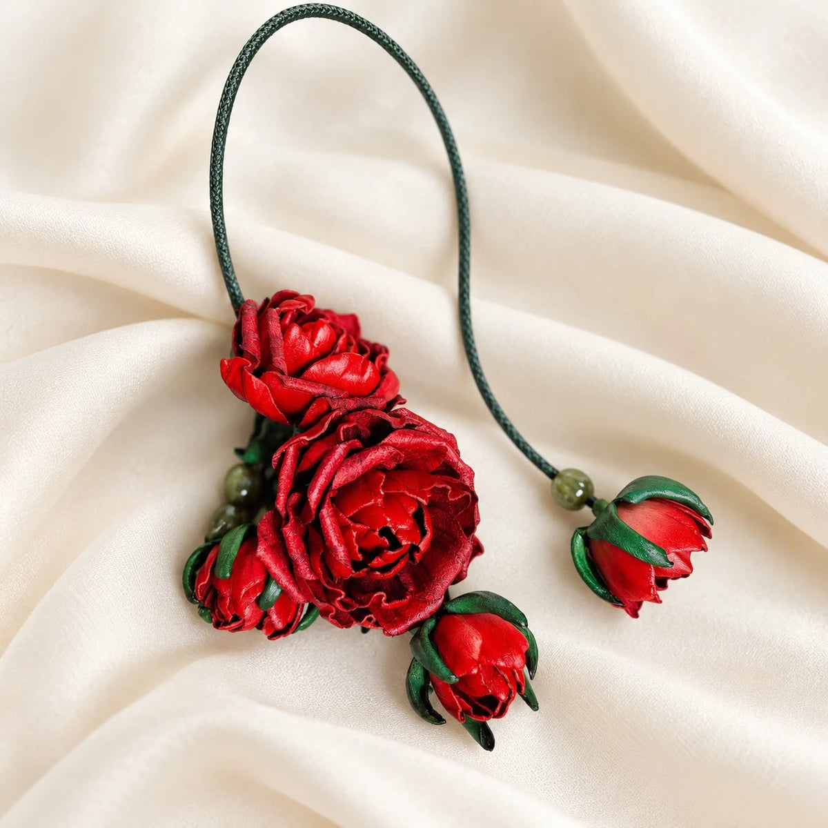 Crimson red sculpted leather rose bag charm with layered blooms, smaller rosebuds, green floral accents, and a dark green hanging cord on ivory fabric.