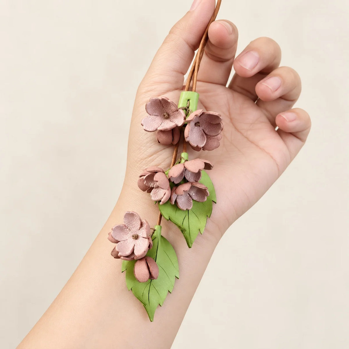 Cherry Blossom Cascade bag charm displayed against a hand and wrist, showing curved pink petals, two veined green leaves, and a slender brown cord.