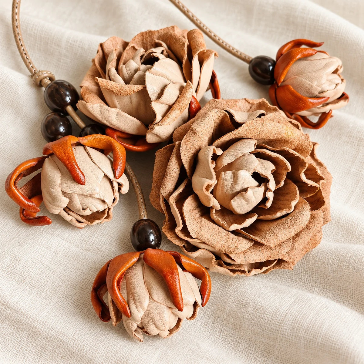 Beige handcrafted leather rose charm arranged on cream linen with two sculpted blossoms, orange-tipped buds, dark beads, and a tan leather cord.