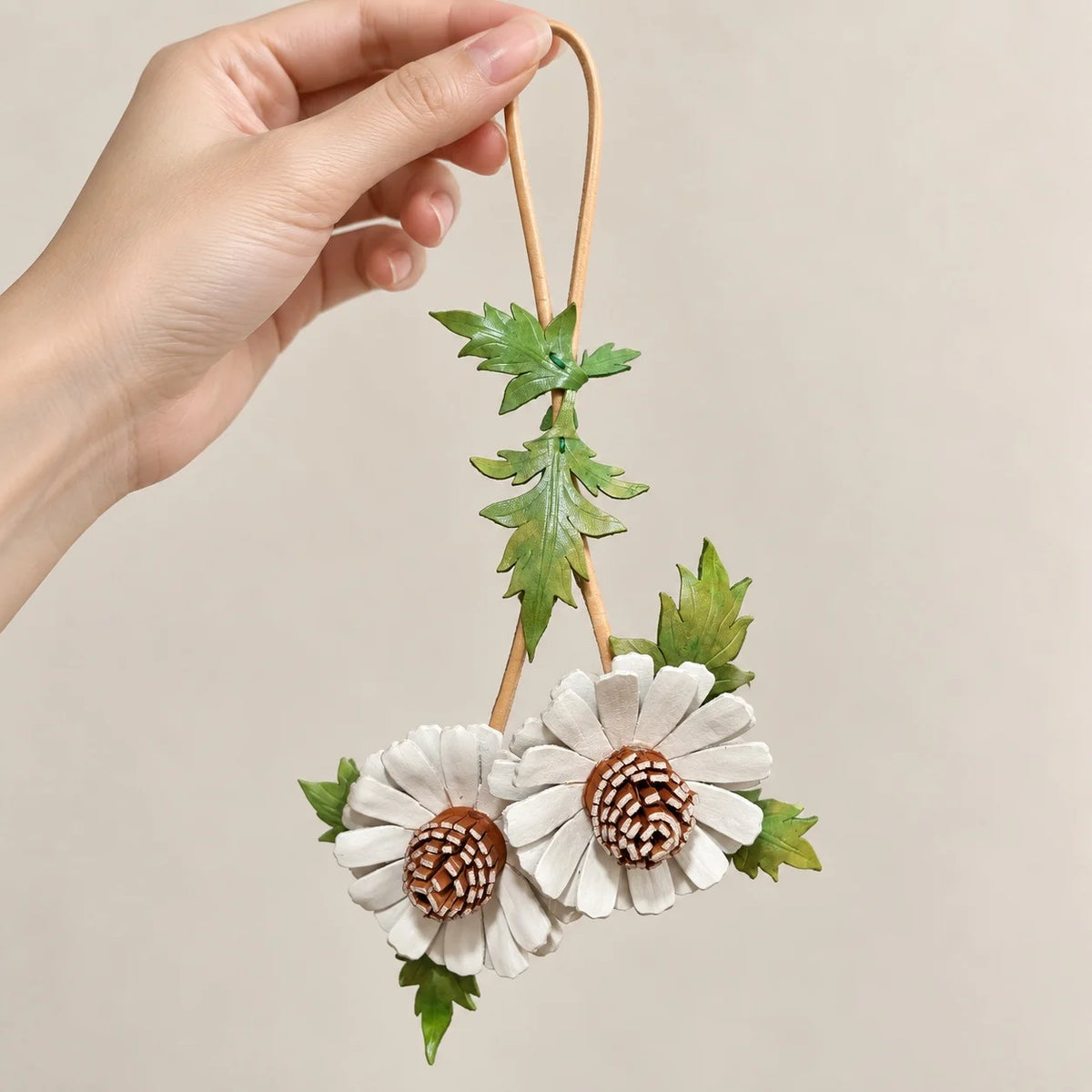 Hand holding the tan loop of a white double daisy leather bag charm, showing its full floral design and trailing green leaves.