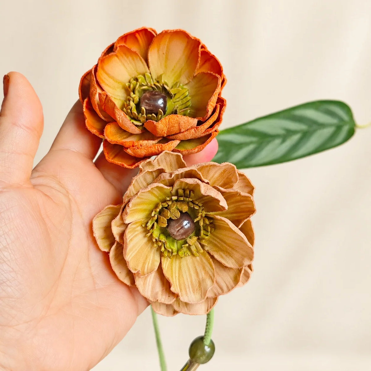 Hand holding an orange and sand double flower leather bag charm, showing layered petals, brown centers and natural flower scale.
