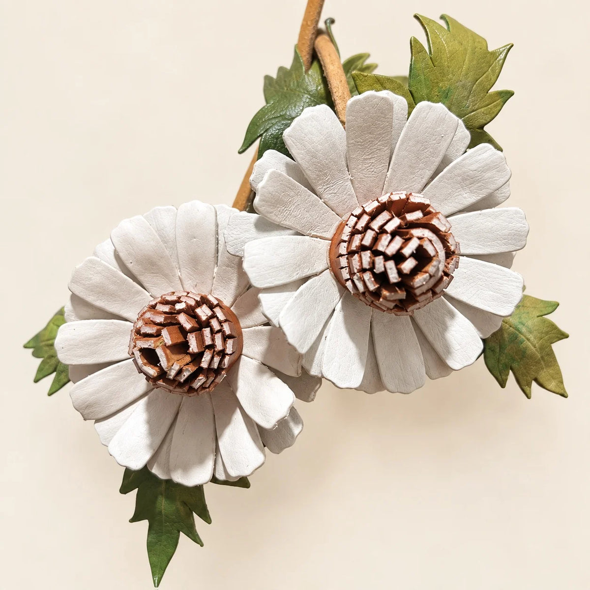 Close-up of two white leather daisies showing dimensional petals, irregular brown leather strip centers and green leaf details.