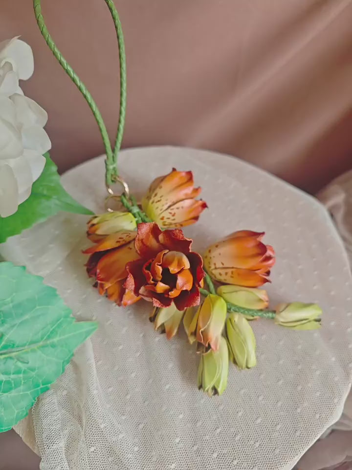Amber Lily Cascade leather bag charm shown by hand, revealing sculpted orange flowers, green buds and gold-tone clasp.