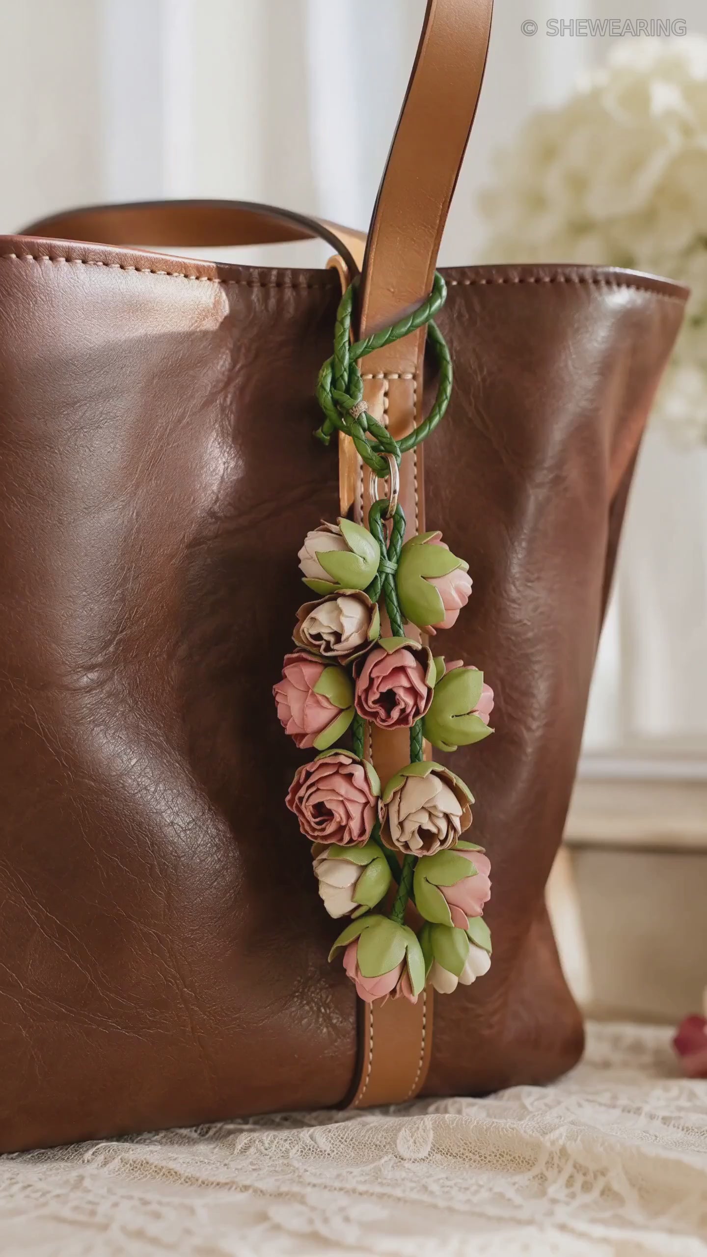 Product video of SHEWEARING Blush Bud Cascade Sculpted Leather Floral Bag Charm, showing pink and cream leather rosebuds, green braided cords and natural movement on a cognac tote.