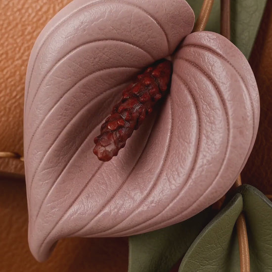 Product video of SHEWEARING Blush Anthurium Cascade Leather Bag Charm, showing two blush pink leather anthurium blooms, burgundy spadices, olive-green leaves and tan cords on a cognac handbag.