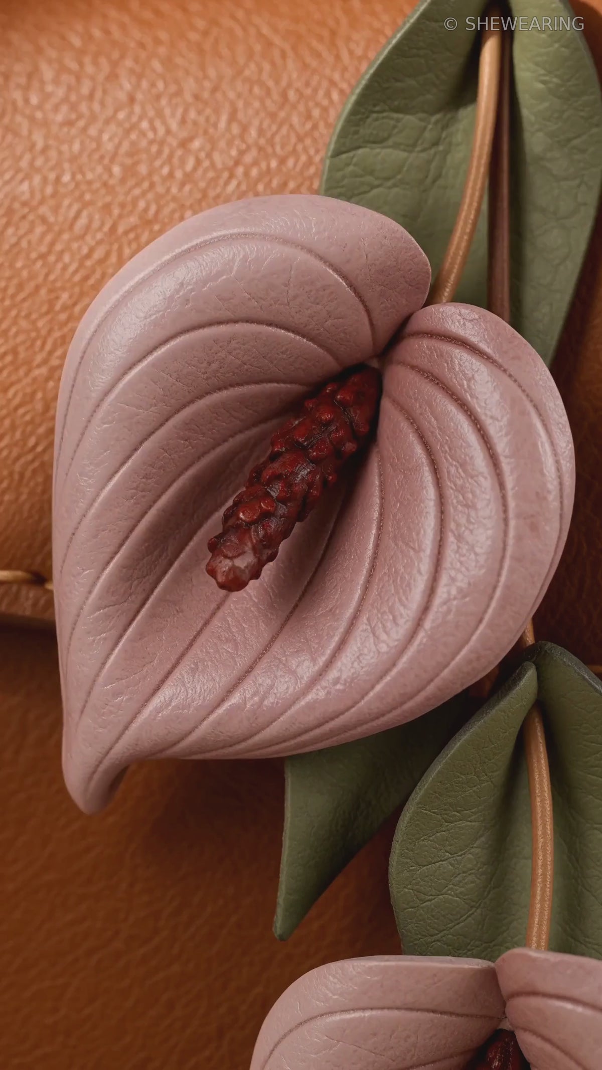 Product video of SHEWEARING Blush Anthurium Cascade Leather Bag Charm, showing two blush pink leather anthurium blooms, burgundy spadices, olive-green leaves and tan cords on a cognac handbag.