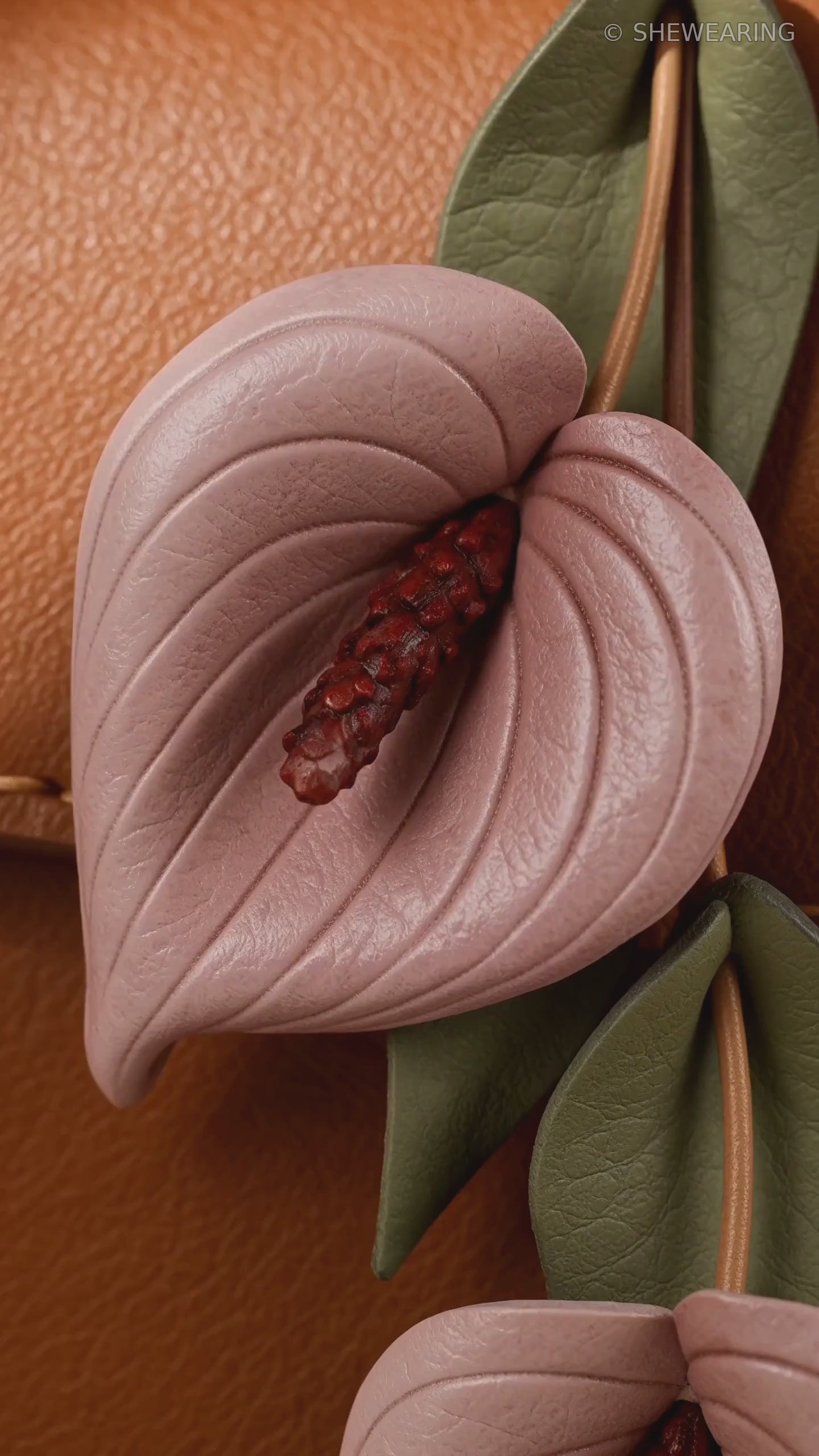 Product video of SHEWEARING Blush Anthurium Cascade Leather Bag Charm, showing two blush pink leather anthurium blooms, burgundy spadices, olive-green leaves and tan cords on a cognac handbag.