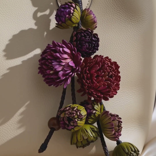 Product video of the SHEWEARING Vineyard Bloom Sculpted Leather Floral Bag Charm, featuring layered amethyst, plum and burgundy leather blossoms, olive-green buds, braided leather cords and wooden bead accents. Shows natural movement and elegant styling on a cream tote handbag.