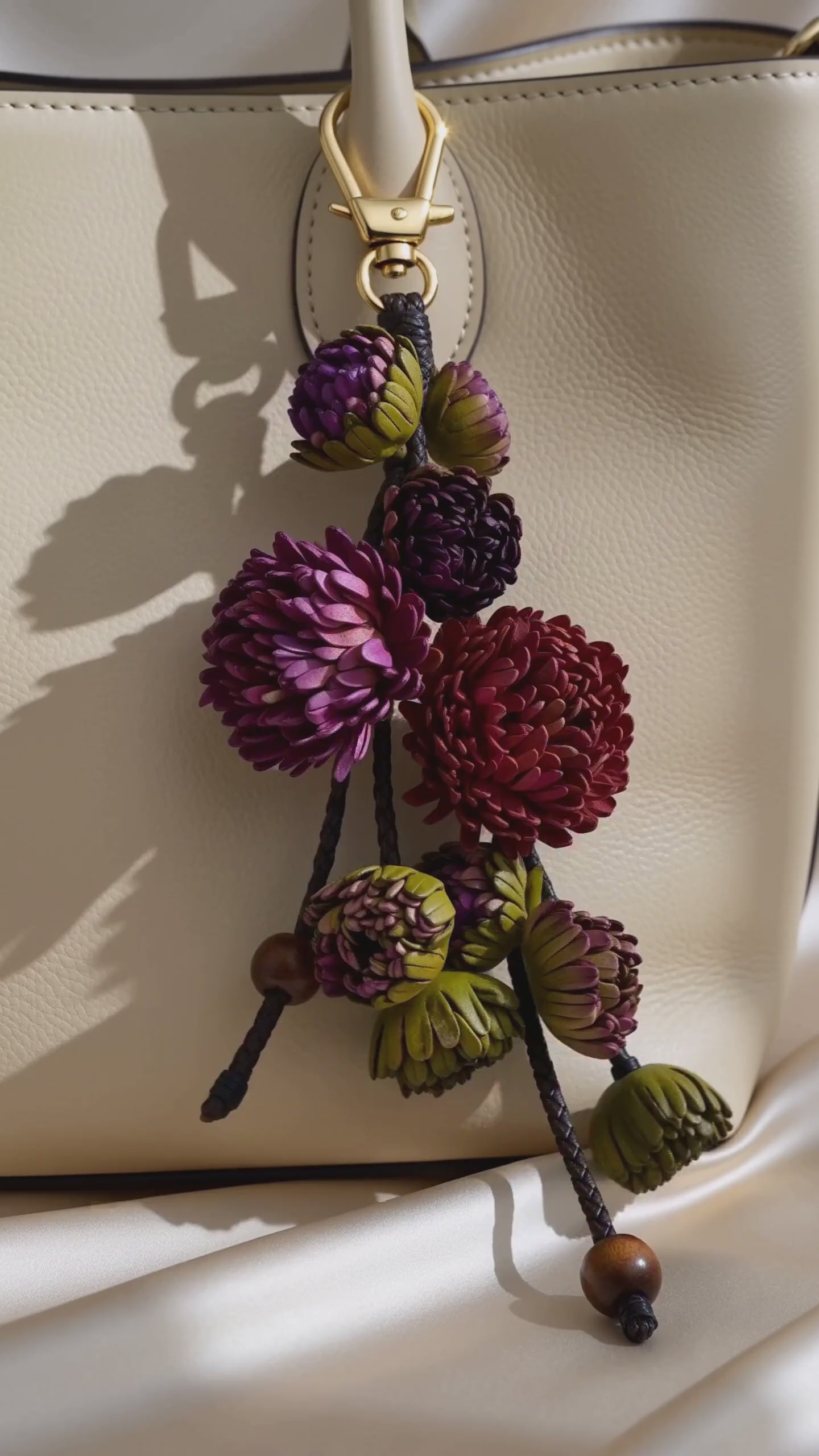 Product video of the SHEWEARING Vineyard Bloom Sculpted Leather Floral Bag Charm, featuring layered amethyst, plum and burgundy leather blossoms, olive-green buds, braided leather cords and wooden bead accents. Shows natural movement and elegant styling on a cream tote handbag.