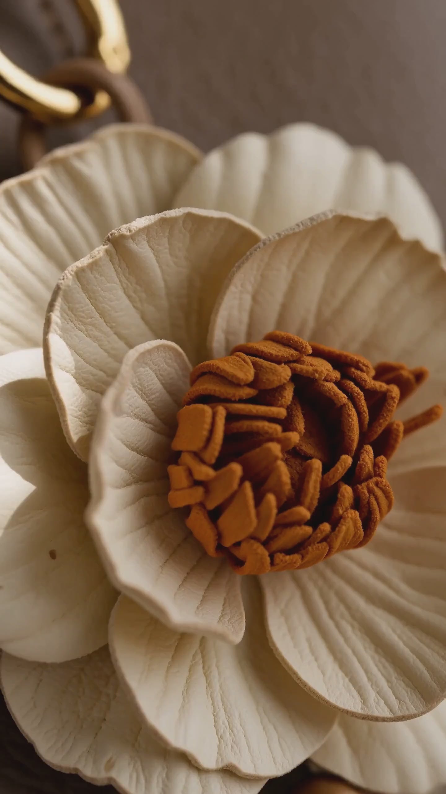 Product video of the SHEWEARING Camellia Grace Sculpted Leather Floral Bag Charm, featuring layered ivory camellia petals, a warm textured center, green leather leaf, dangling flower bud, natural bead accents, and gold-tone hardware, styled on a warm taupe handbag.