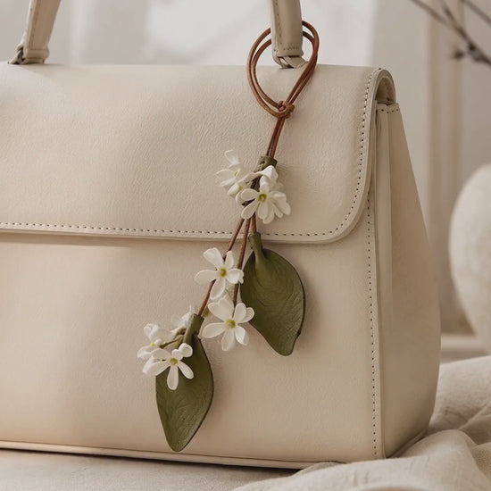 SHEWEARING White Star Jasmine leather bag charm video showing delicate white flowers, two olive-green leaves and brown cords on a cream handbag.