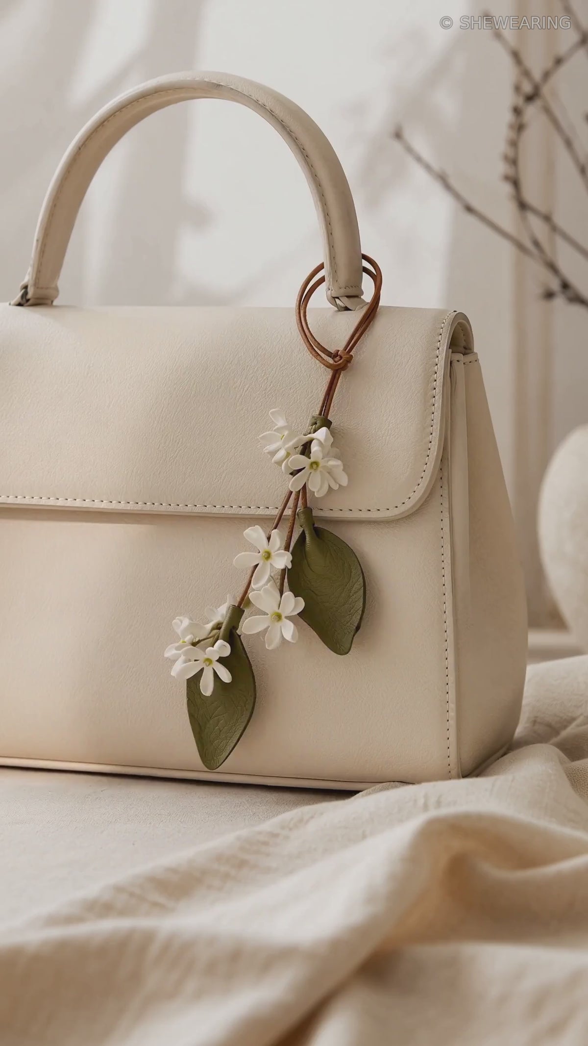 SHEWEARING White Star Jasmine leather bag charm video showing delicate white flowers, two olive-green leaves and brown cords on a cream handbag.