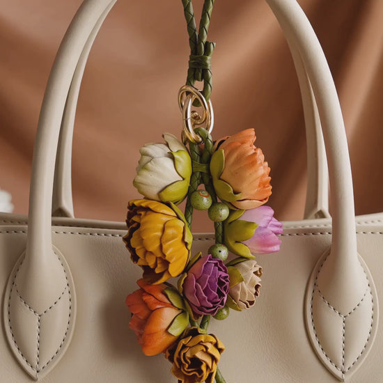 Product video of the SHEWEARING Primavera Bloom Cascade Sculpted Leather Floral Bag Charm, showing a cascading arrangement of multicolor leather blossoms and buds with green braided cords and gold-tone hardware, styled on a cream tote handbag with soft natural movement.