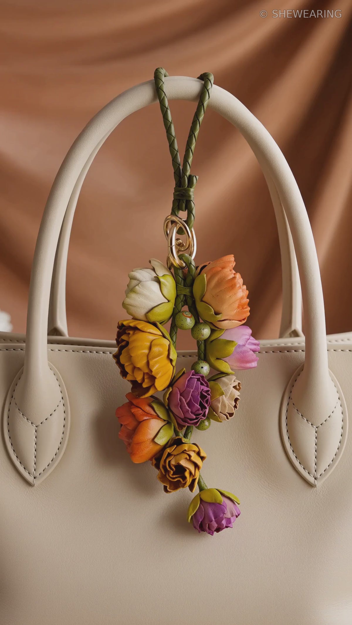 Product video of the SHEWEARING Primavera Bloom Cascade Sculpted Leather Floral Bag Charm, showing a cascading arrangement of multicolor leather blossoms and buds with green braided cords and gold-tone hardware, styled on a cream tote handbag with soft natural movement.