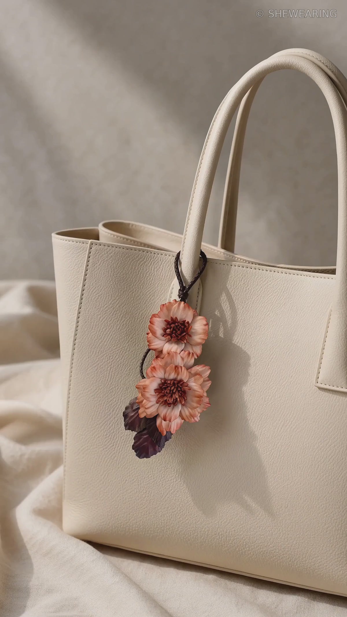 Product video of SHEWEARING Peony Reverie Sculptural Leather Bag Charm, featuring two coral leather peony blooms, textured centers, burgundy leaves and gentle movement on a cream tote.