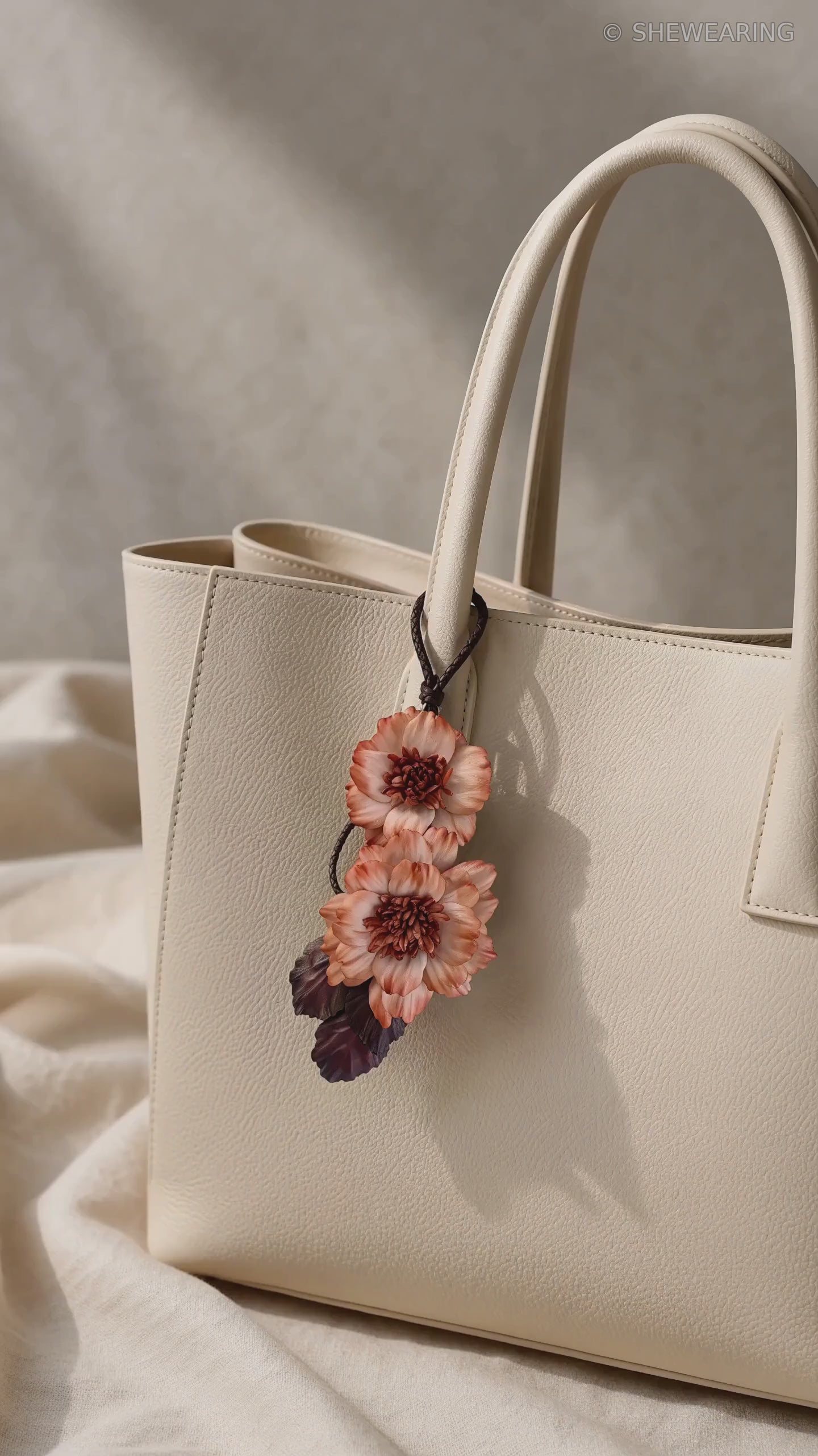 Product video of SHEWEARING Peony Reverie Sculptural Leather Bag Charm, featuring two coral leather peony blooms, textured centers, burgundy leaves and gentle movement on a cream tote.